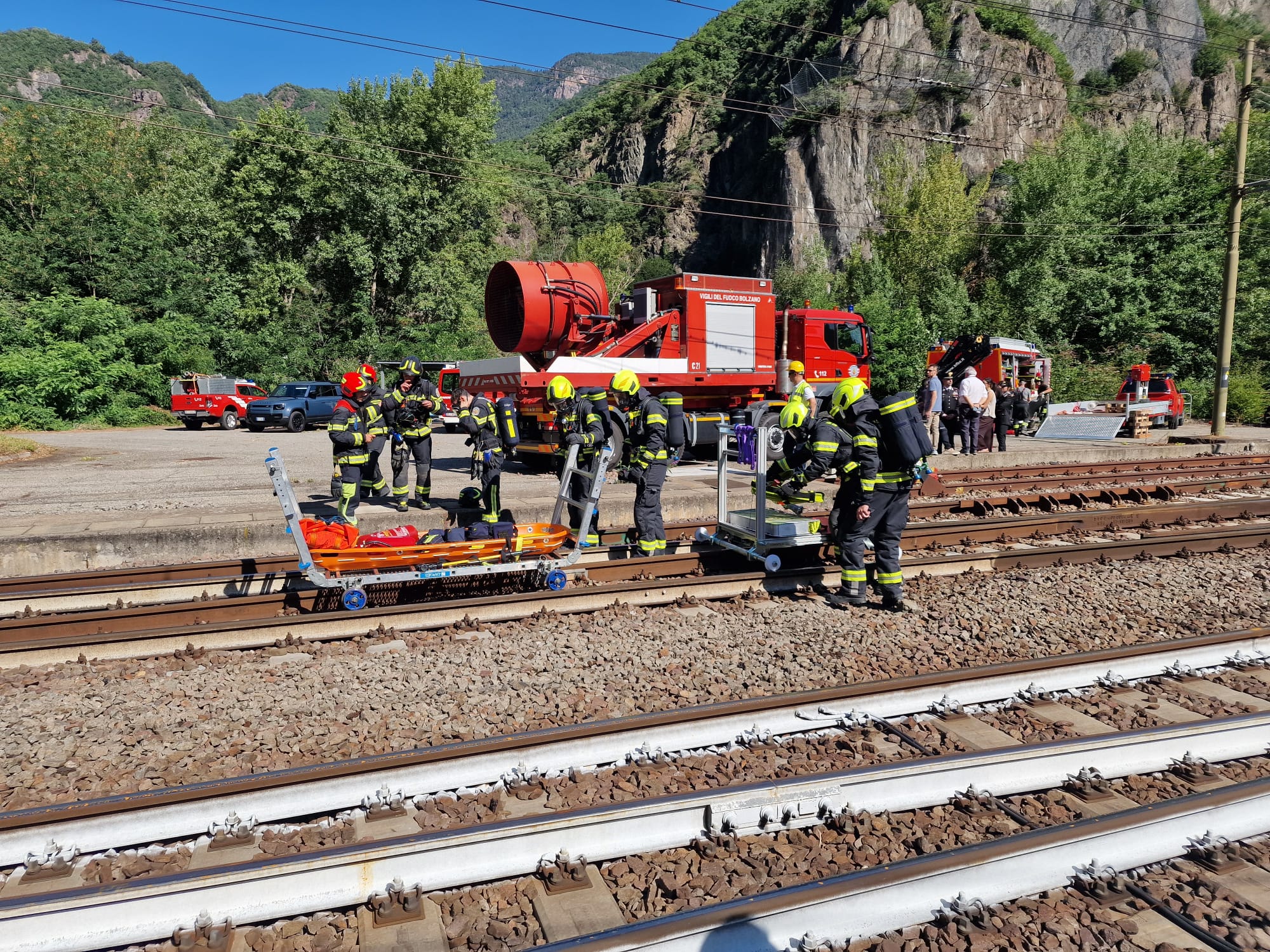 Firefighters in gear stand around a red fire truck and rescue equipment beside train tracks, with green hills in the background of a sunny scene.
